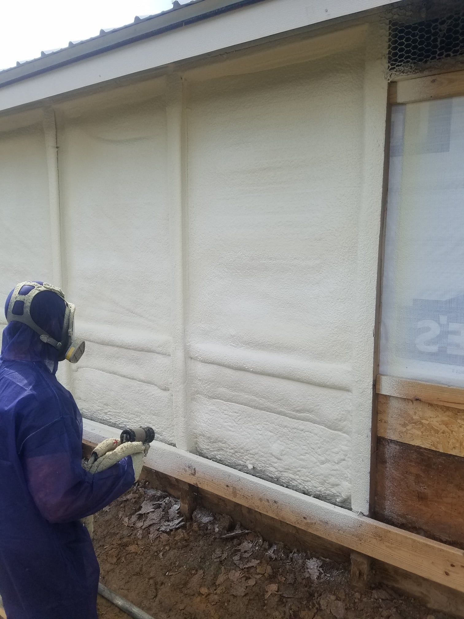Insulation technician in protective gear spraying foam along an interior wall